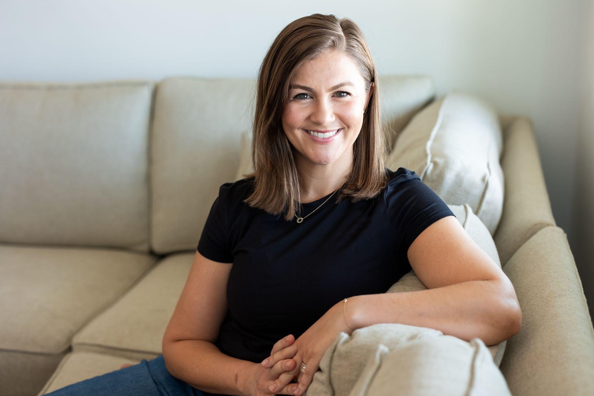 Woman with short brown hair smiling, seated on a beige couch in a relaxed, indoor setting.